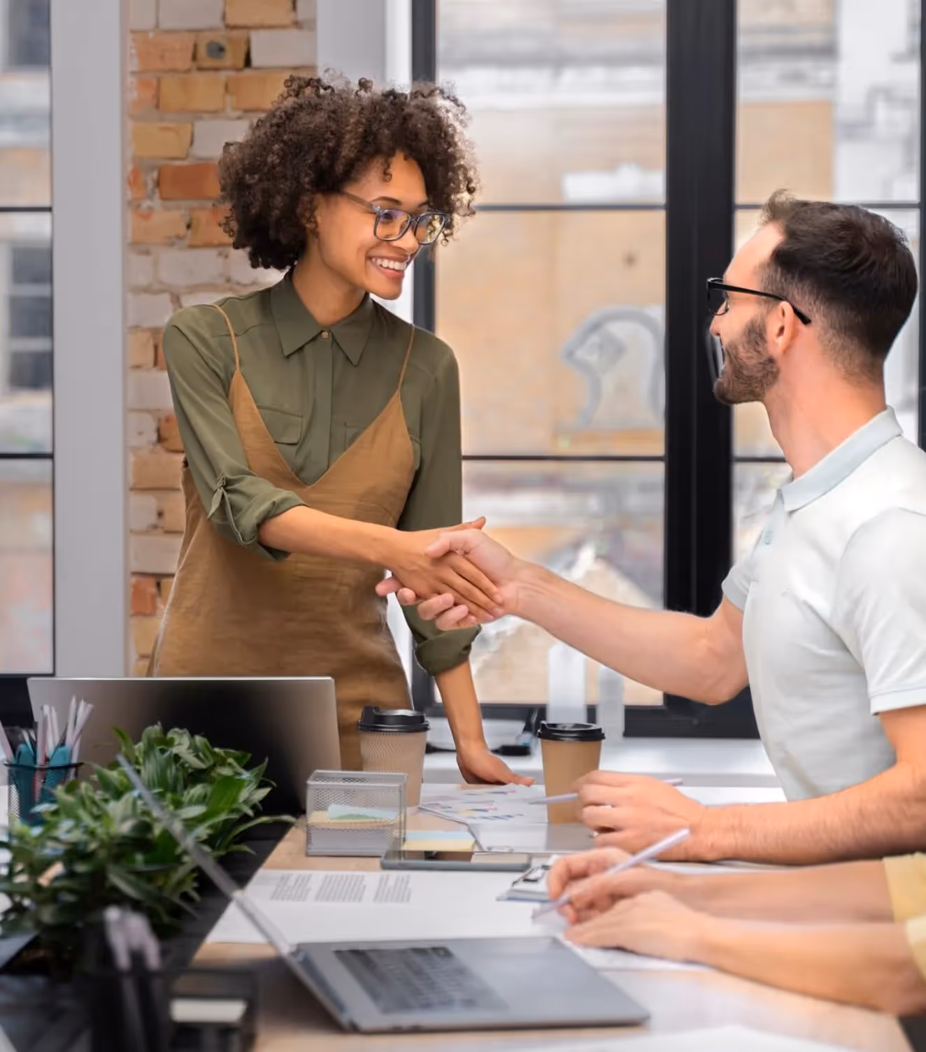 Smiling woman and man shaking hands across a table with laptops and coffee cups in a modern office.