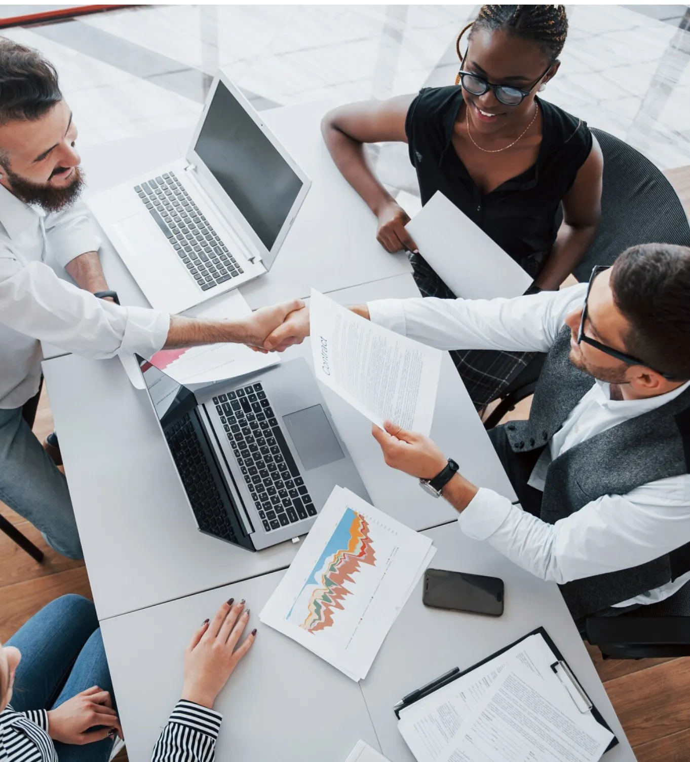 Two men shaking hands over a table with laptops and documents, while two colleagues observe.