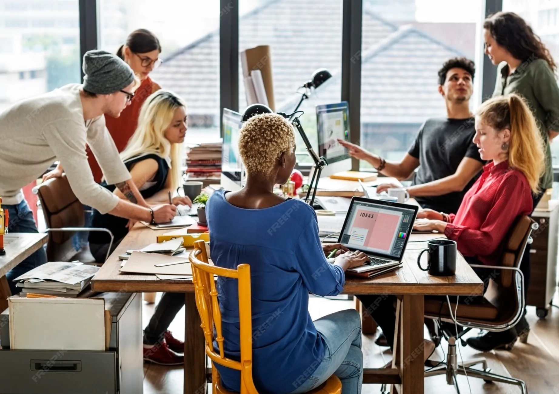 Diverse group of six young professionals collaborating around a table in a bright office workspace with laptops and documents.