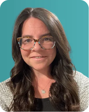 Smiling woman with long dark wavy hair, wearing glasses, a gray sweater, and a necklace, against a teal background.