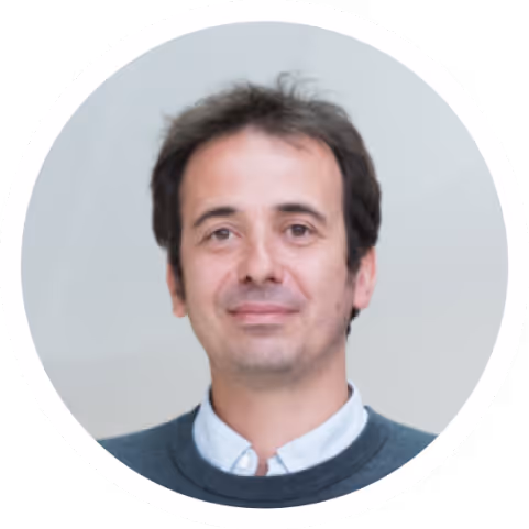 Portrait of a man with short dark hair wearing a white collared shirt and dark sweater, smiling softly against a neutral background.