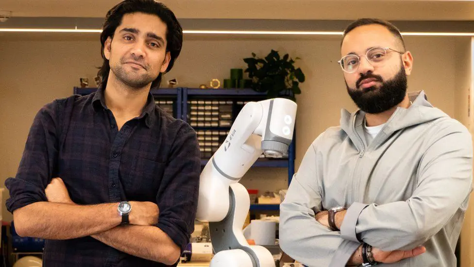 Two men with arms crossed standing beside a white robotic arm in a workshop setting.
