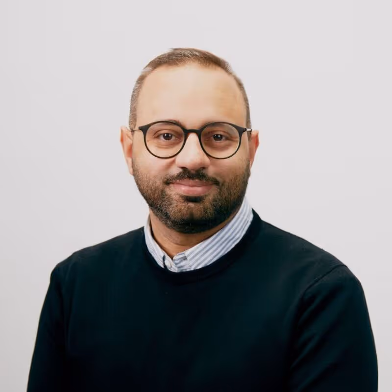 Man with glasses and beard wearing a black sweater over a striped collared shirt against a light background.