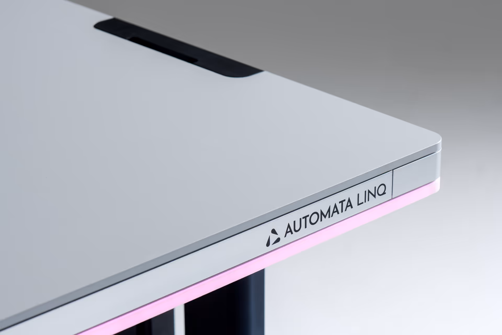 Close-up of the corner of a sleek gray desk with a subtle triangular logo and the word Automata Linq, illuminated by a soft pink LED strip underneath.