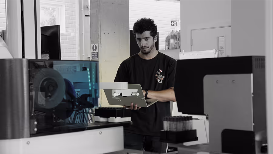 Man in a lab holding a laptop, standing among laboratory machines and sample tubes.