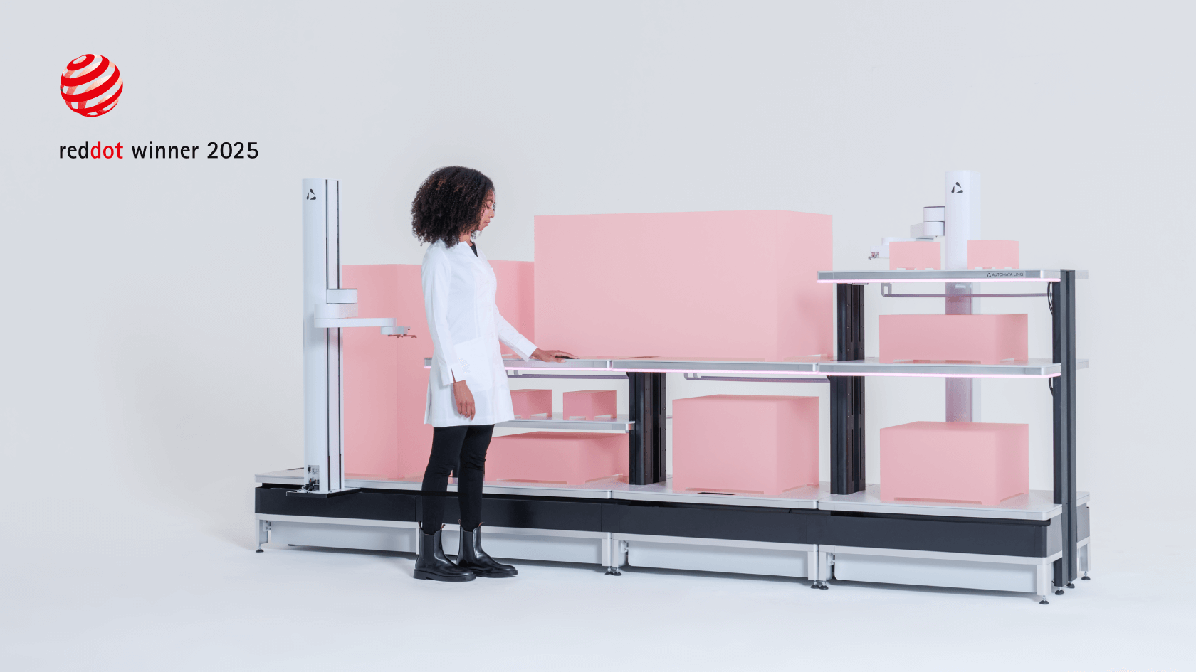 Woman in a white lab coat standing beside a modern automated shelving system with pink boxes stacked on it and a Red Dot winner 2025 logo in the background.