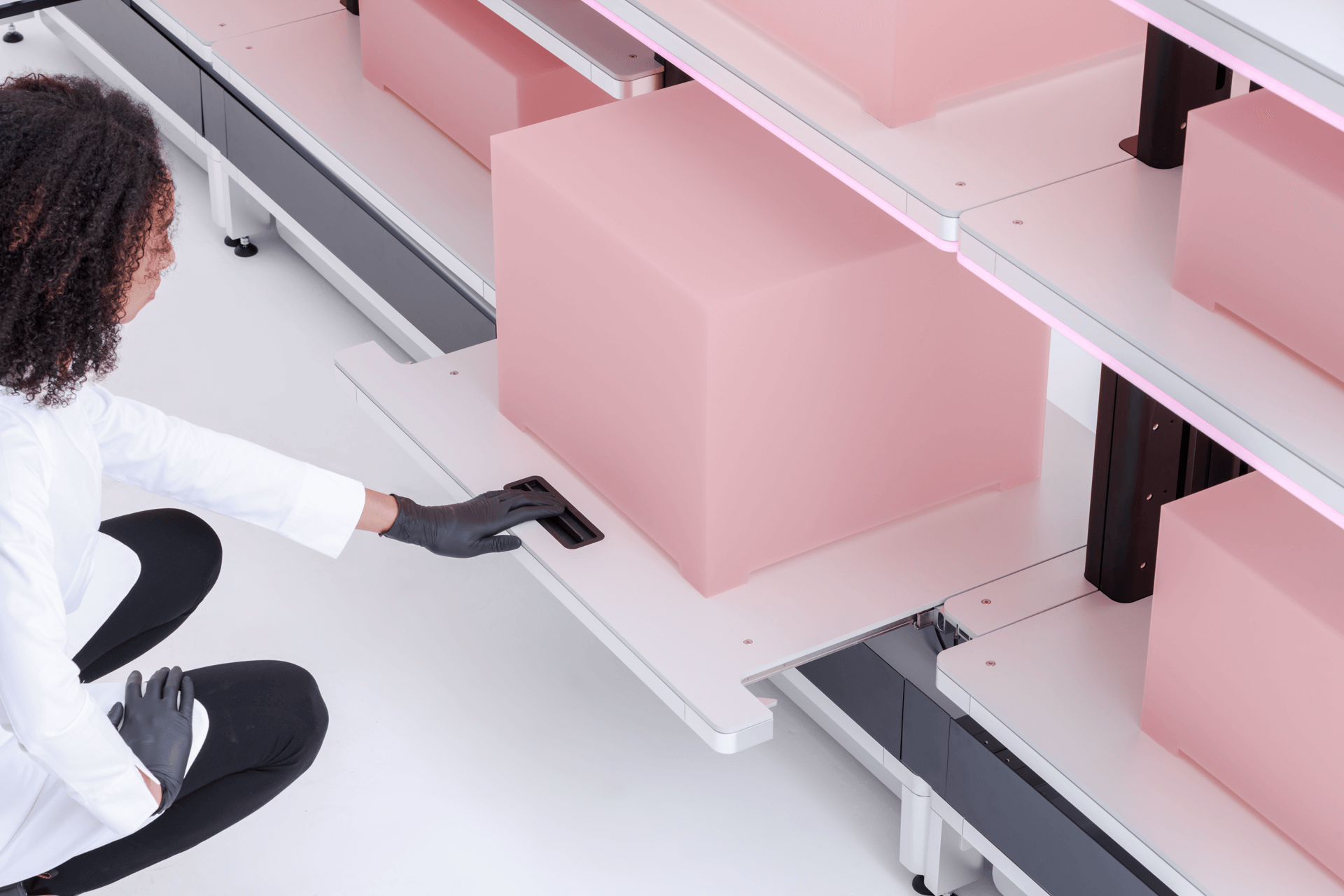 Person wearing black gloves and white coat sliding out a white shelf holding a large pink box in a clean, modern storage system.