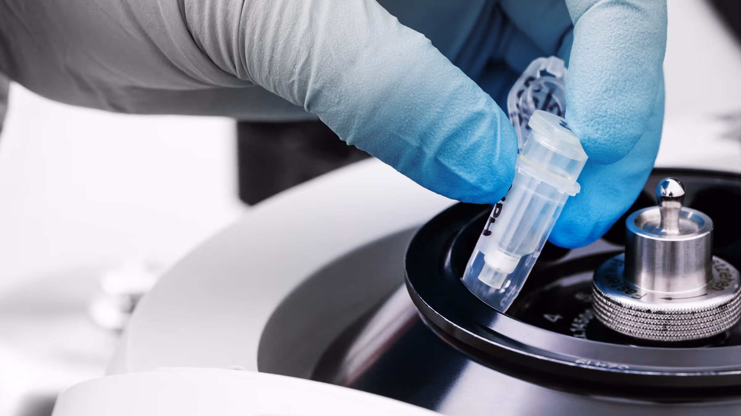 Gloved hand placing a small vial into a centrifuge rotor in a laboratory setting.