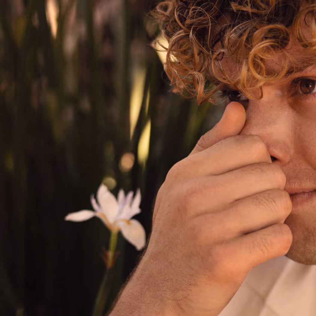 Close-up of a person’s face and hand suggesting an allergy, with blurred greenery and a flower in the background.