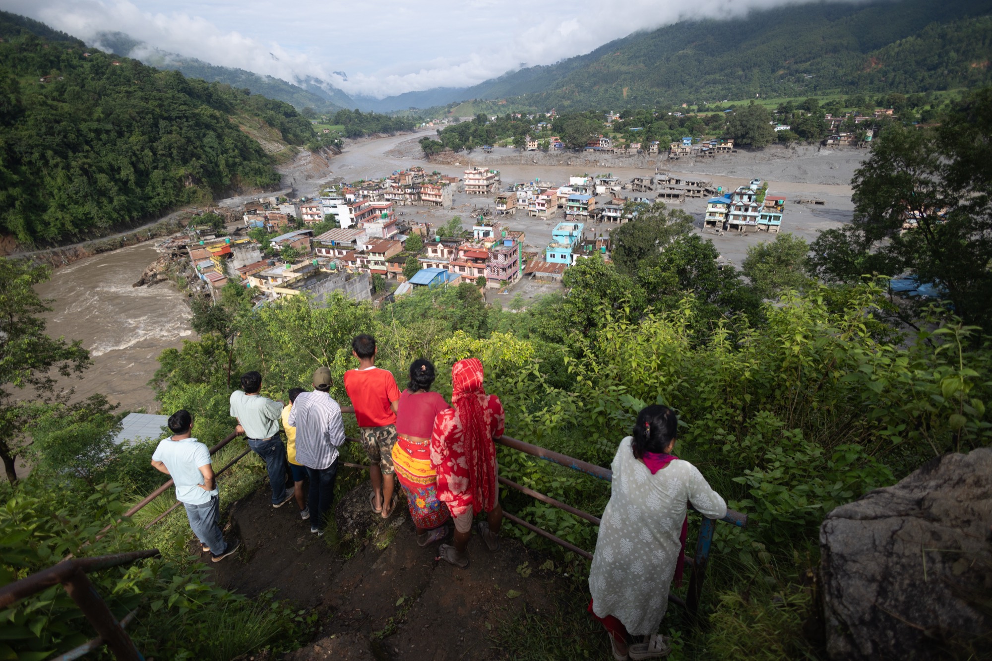 People look out over a Nepalese town damaged by the 2026 floods, with debris and floodwater filling the valley below.