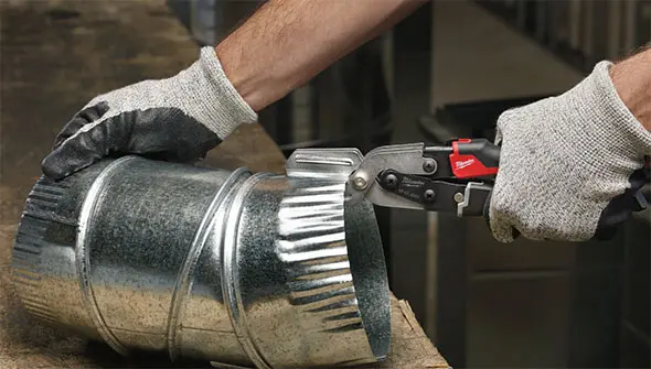 A worker wearing gloves uses red Milwaukee aviation snips to cut a crimped galvanized metal duct pipe.