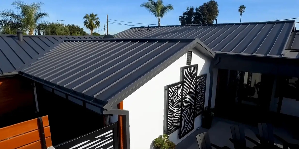 Modern dark gray metal standing seam roof with clean lines on a contemporary white stucco home exterior, palm trees in background.