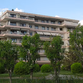 Immeuble résidentiel beige de plusieurs étages avec balcons, entouré d'arbres verts et buissons.