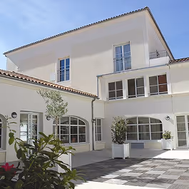 Cour intérieure lumineuse d'un bâtiment moderne avec murs blancs, fenêtres à volets, ciel bleu et plantes en pots.
