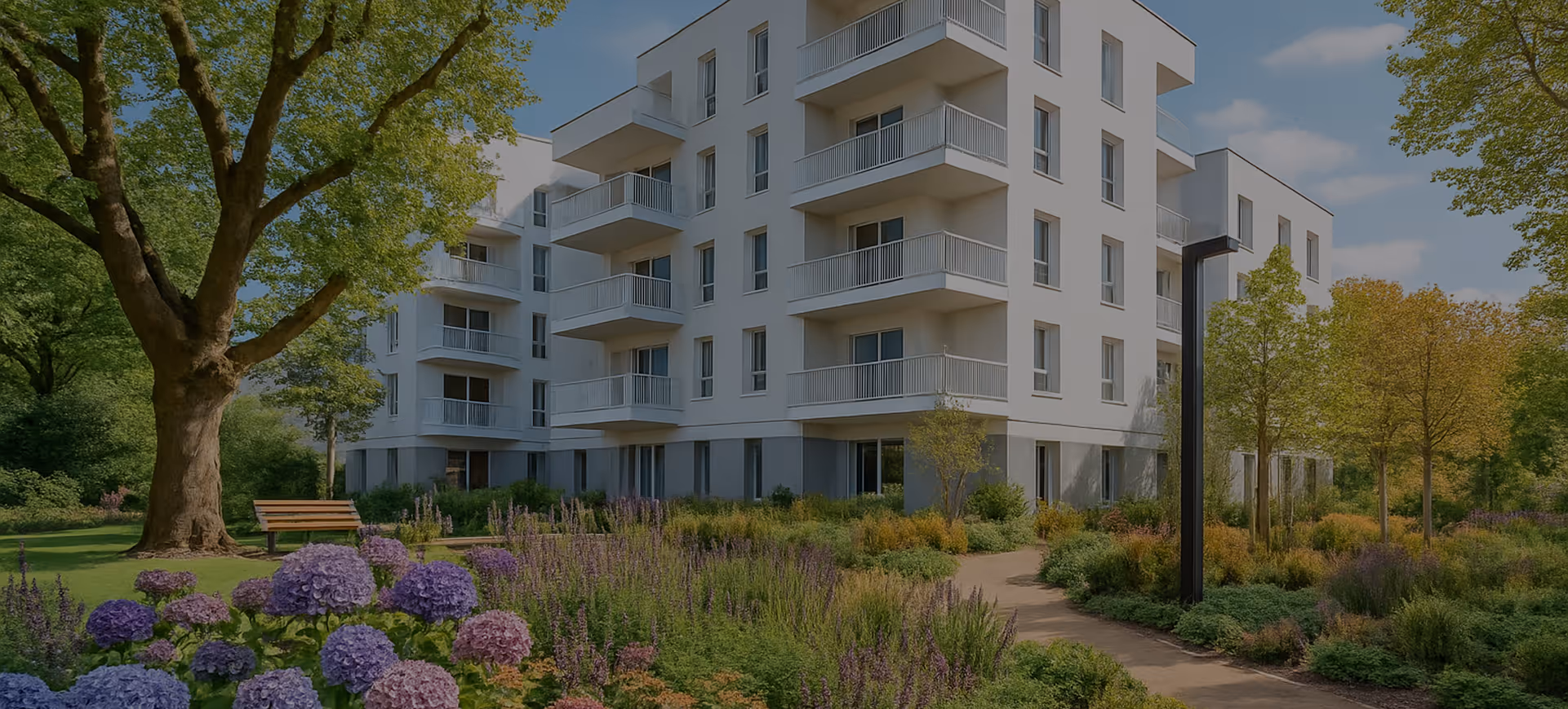Appartement moderne blanc de quatre étages avec balcons, entouré de jardins bien entretenus avec fleurs violettes, arbres et chemin de promenade.