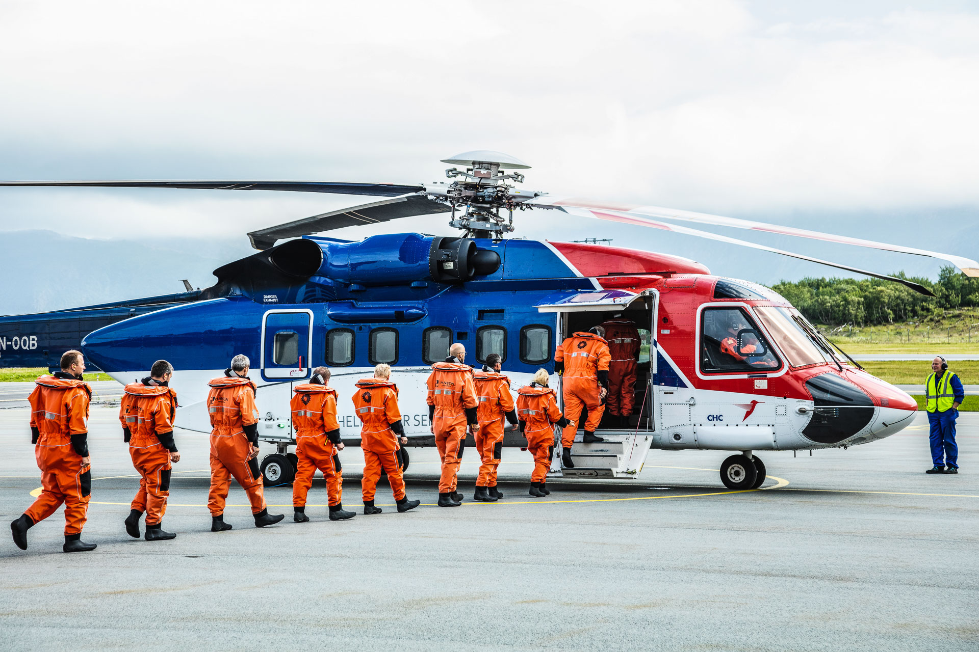 En gruppe personer i oransje vernedrakter går mot en blå og rødfarget helikopter på en flyplass.