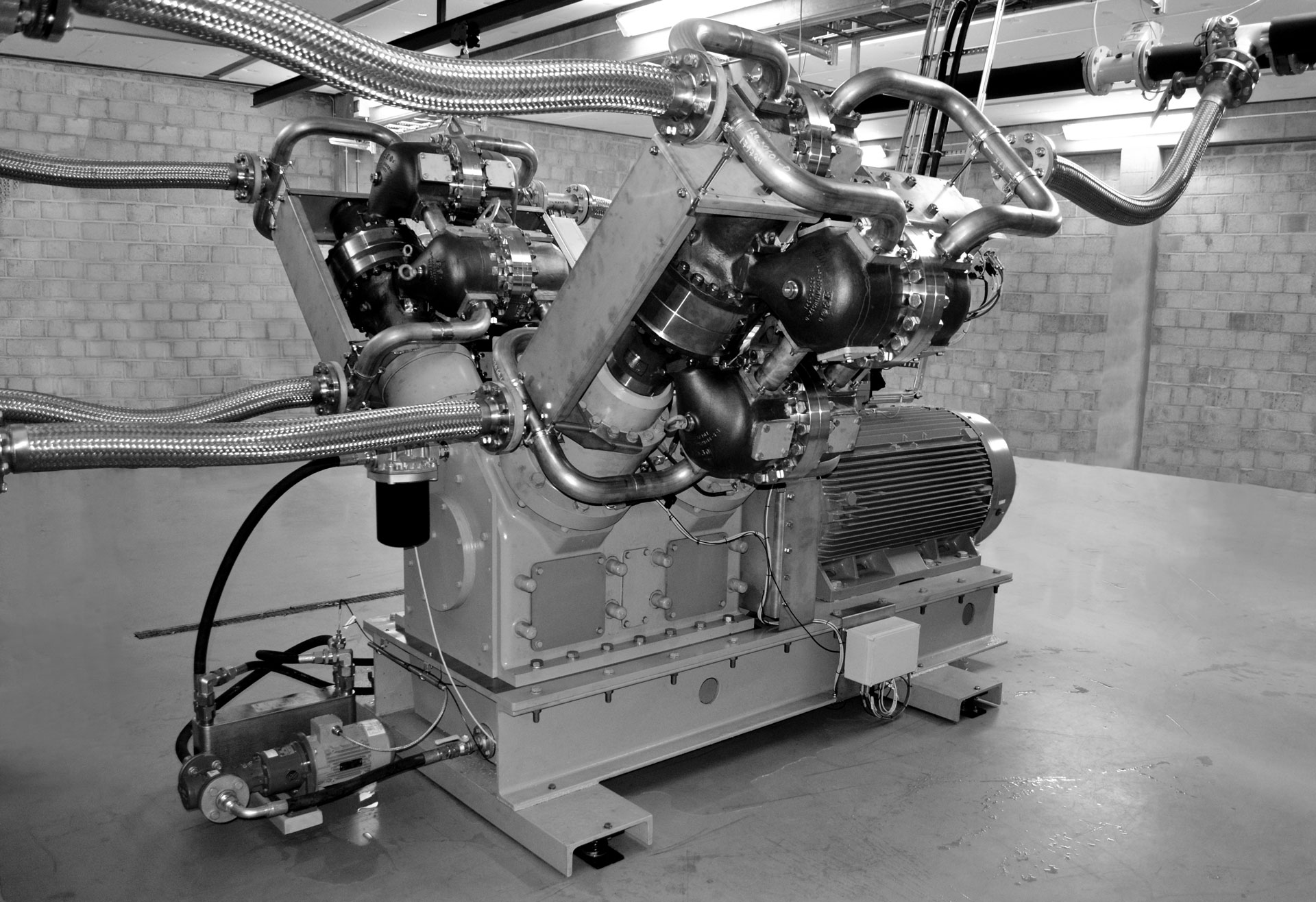 Industriell motor med flere koblede rør og metallslanger i et rom med teglsteinsvegger.