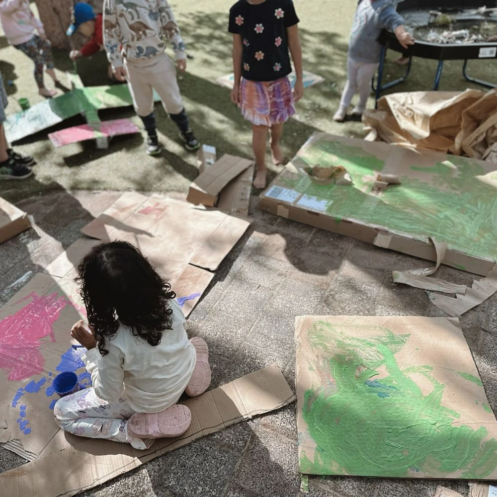 A little girl sitting on the ground painting on cardboard.