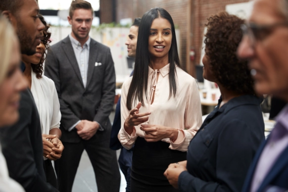 A diverse group of professionals attentively listening to a woman speaking in a business setting.