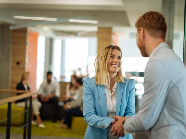 A woman in a blue blazer smiling and shaking hands with a man in a light-colored shirt in an office setting.