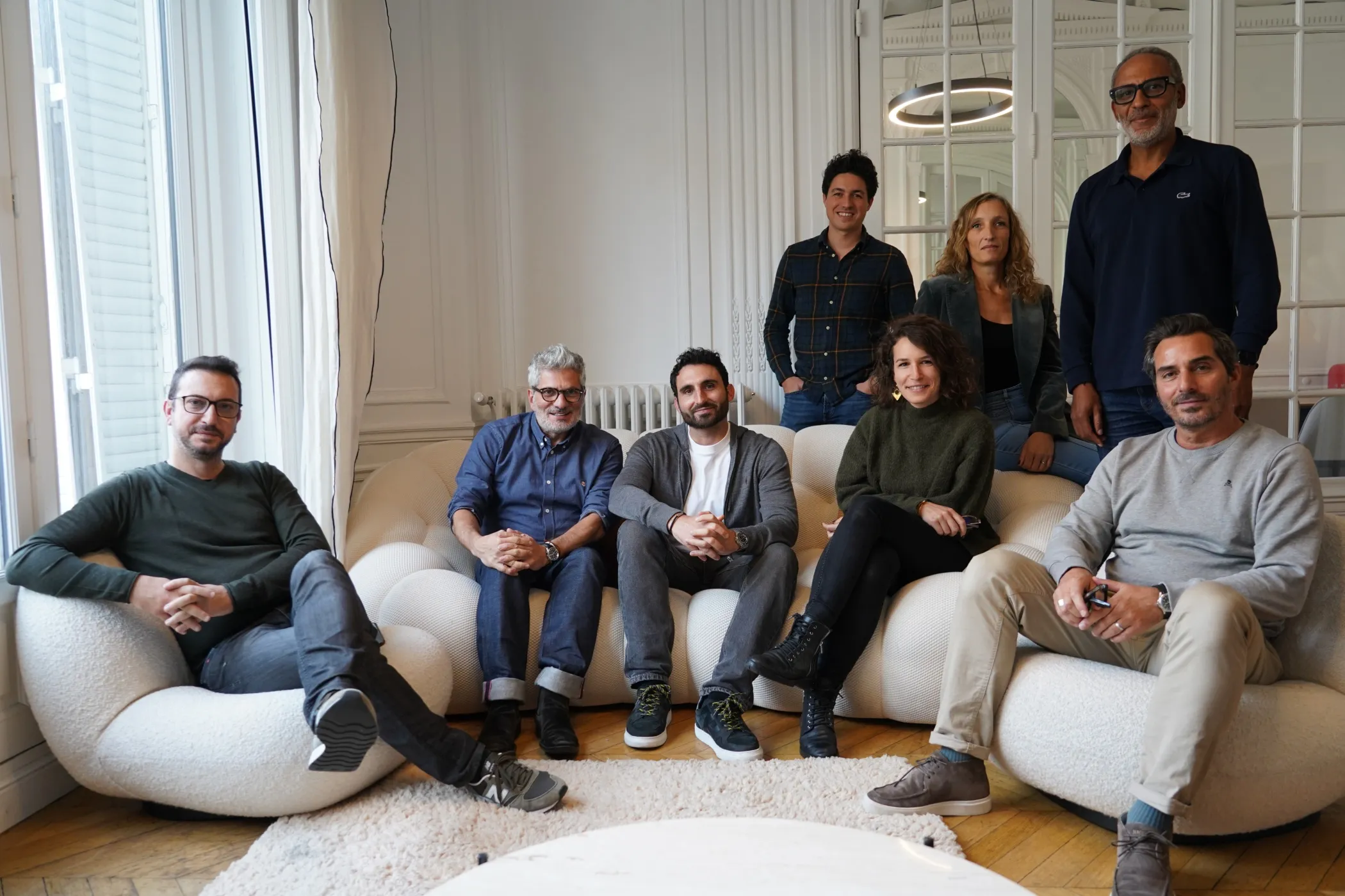 Group of seven people sitting and standing in a stylish living room with white walls and a large window.