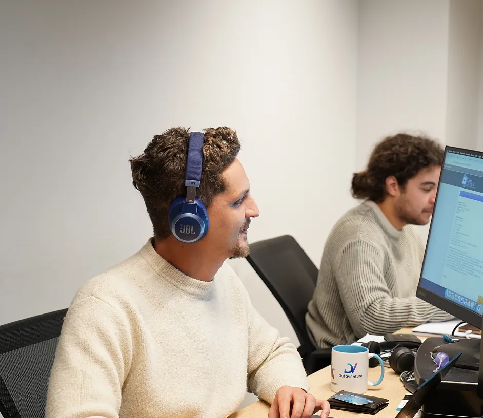 Two men working at desks with computers; the man in front wears blue JBL headphones and a cream sweater, while the man in the back has curly hair and a gray sweater.