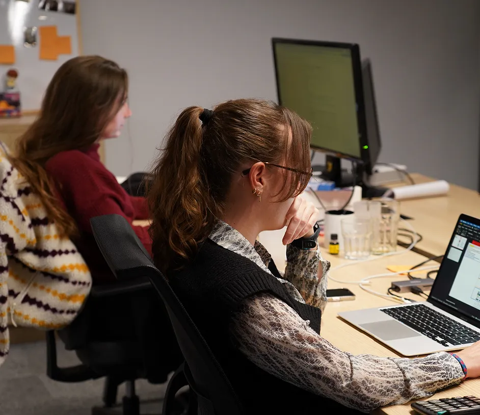 Three women working at desks with laptops and monitors in an office setting.