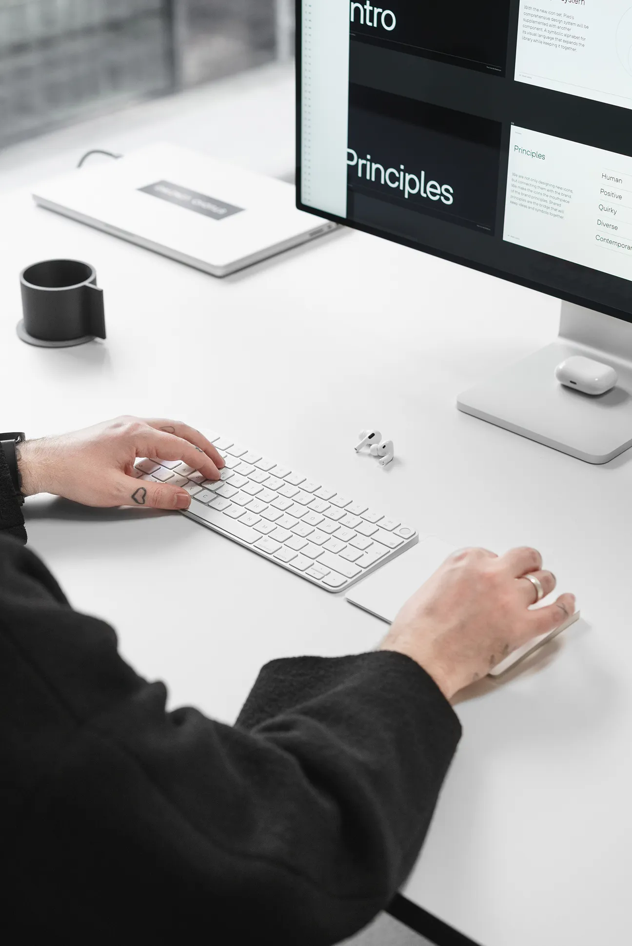 Person using white keyboard and mouse at a clean white desk with a monitor displaying text and wireless earbuds nearby.