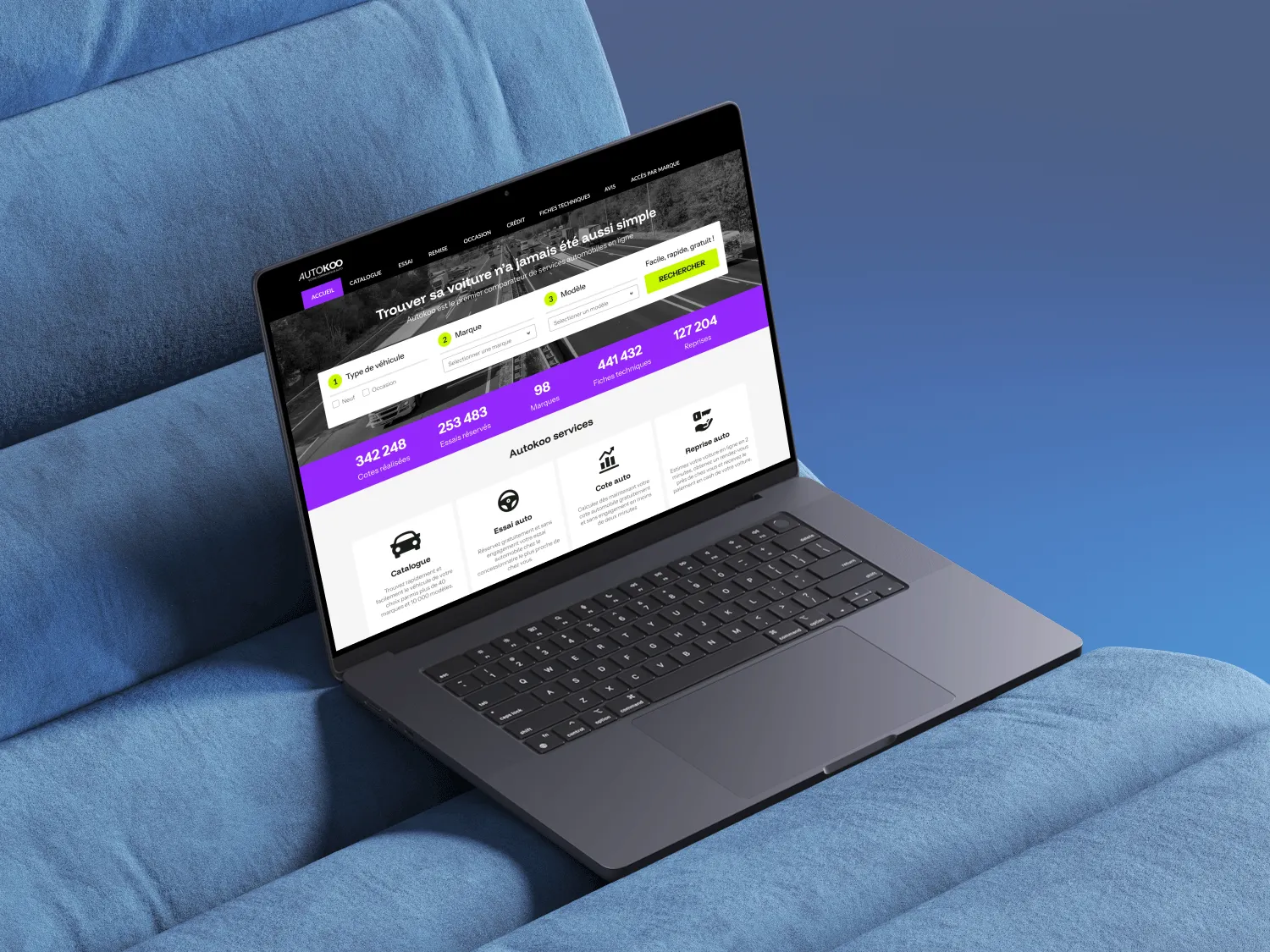 Laptop on blue fabric chair displaying a car comparison and services website with dropdown filters for vehicle type, brand, and model.