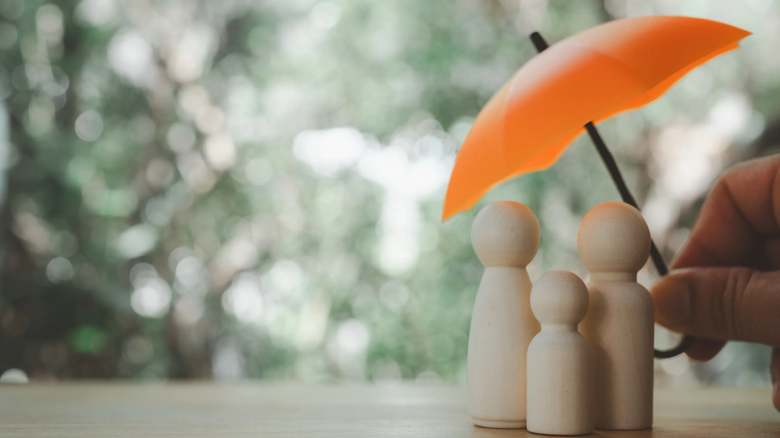 Three wooden figurines representing a family sheltered under an orange umbrella held by a hand.