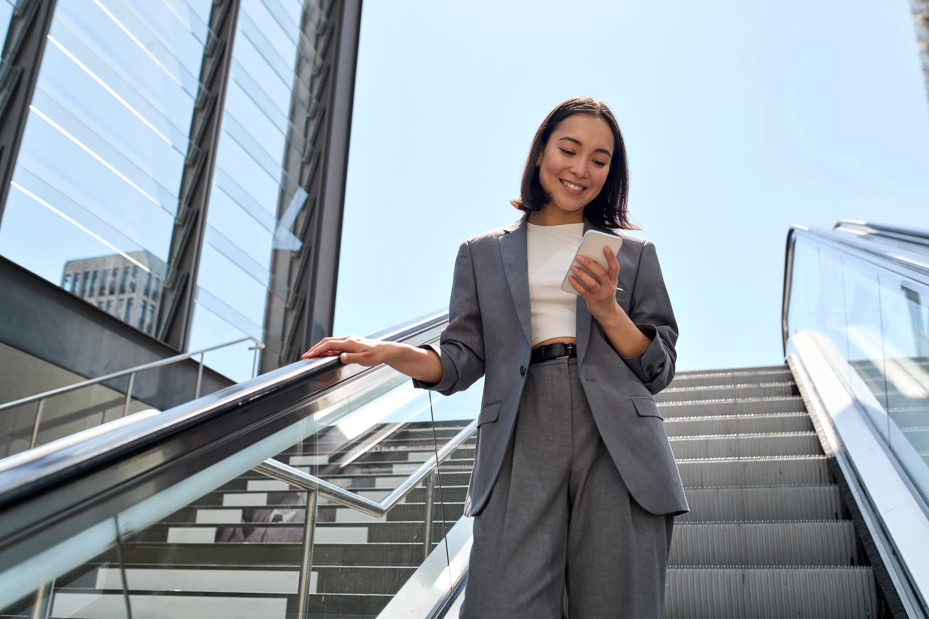 Smiling woman in gray suit riding down an escalator while looking at her smartphone.
