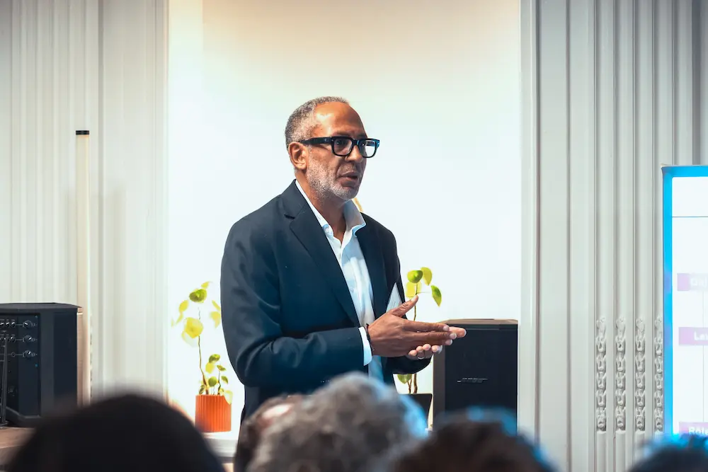 Man wearing glasses and a dark blazer speaking in front of an audience in a well-lit room with plants and a presentation screen.