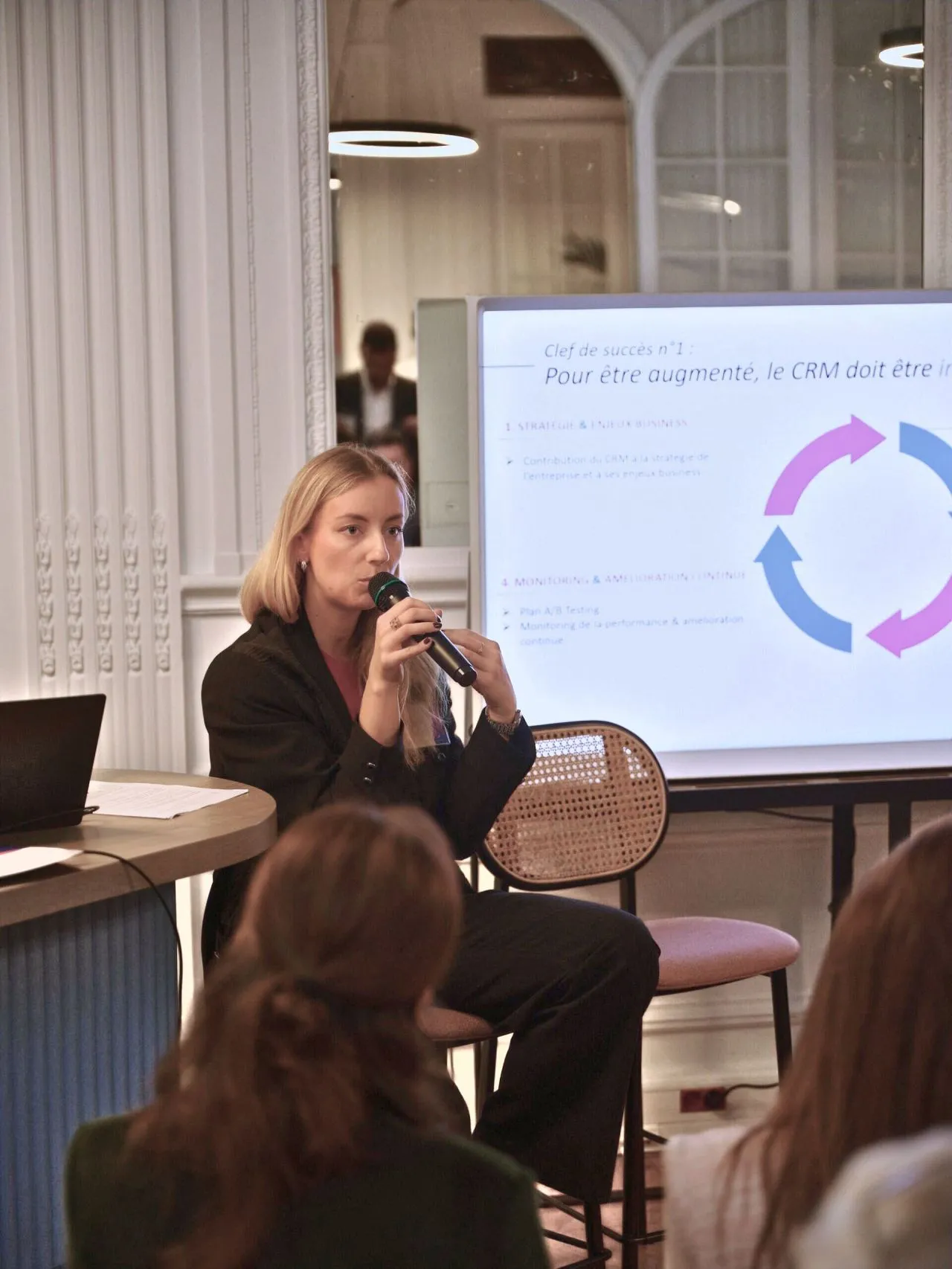 Woman seated and speaking into a microphone during a presentation with a slide about CRM strategy and business visible behind her.