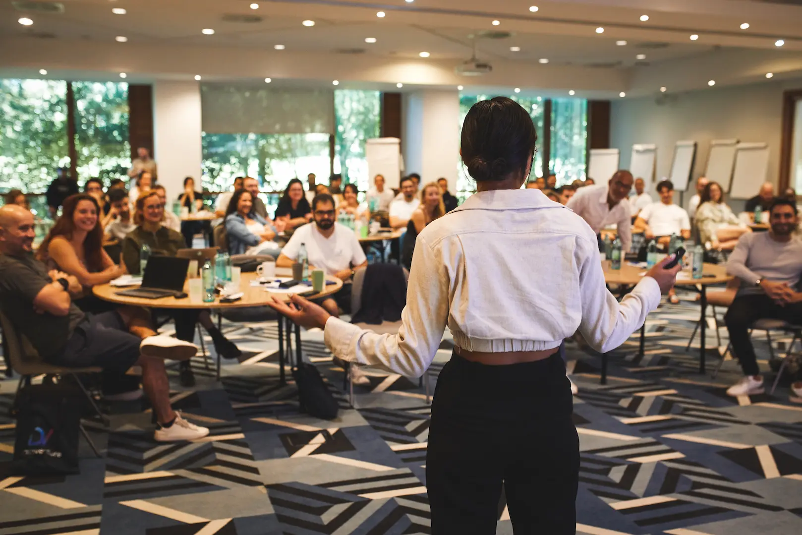 Woman giving a presentation to an attentive audience seated around tables in a conference room.