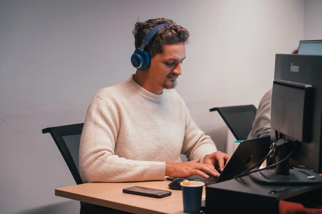 Man wearing headphones and a beige sweater typing on a laptop at a desk with a smartphone and coffee cup.