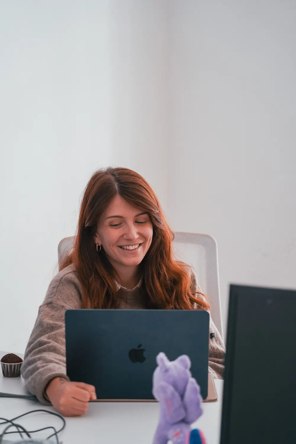 Smiling woman with long brown hair working on a black Apple laptop at a white desk with a purple plush toy in the foreground.