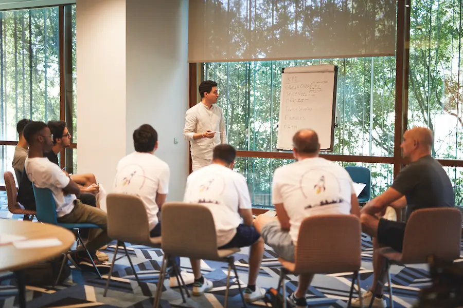 Man standing and presenting to a group of seated people around a flip chart in a bright room with large windows.