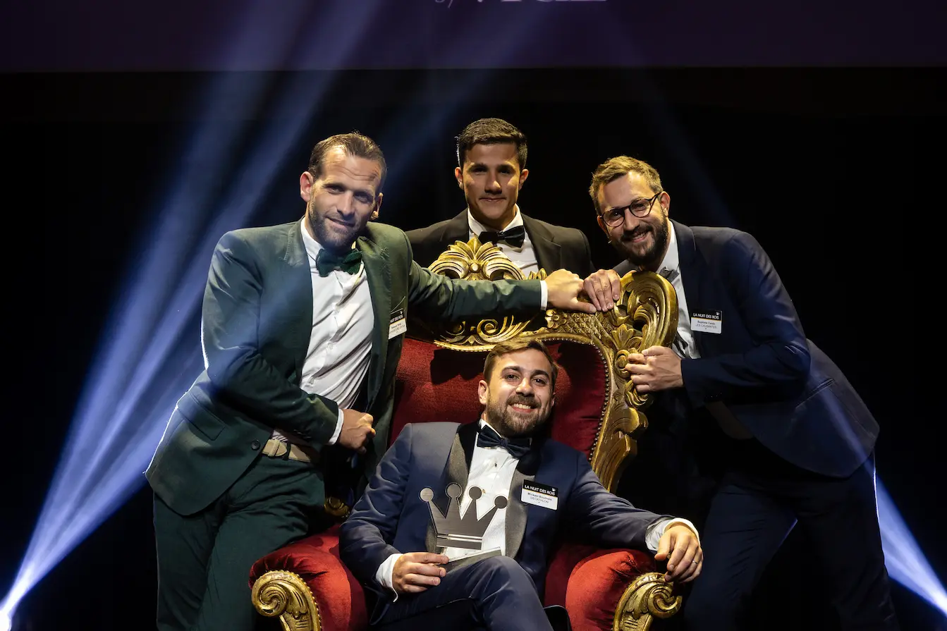 Four men dressed in formal suits posing with a crown-shaped trophy; one seated on an ornate red and gold throne chair.