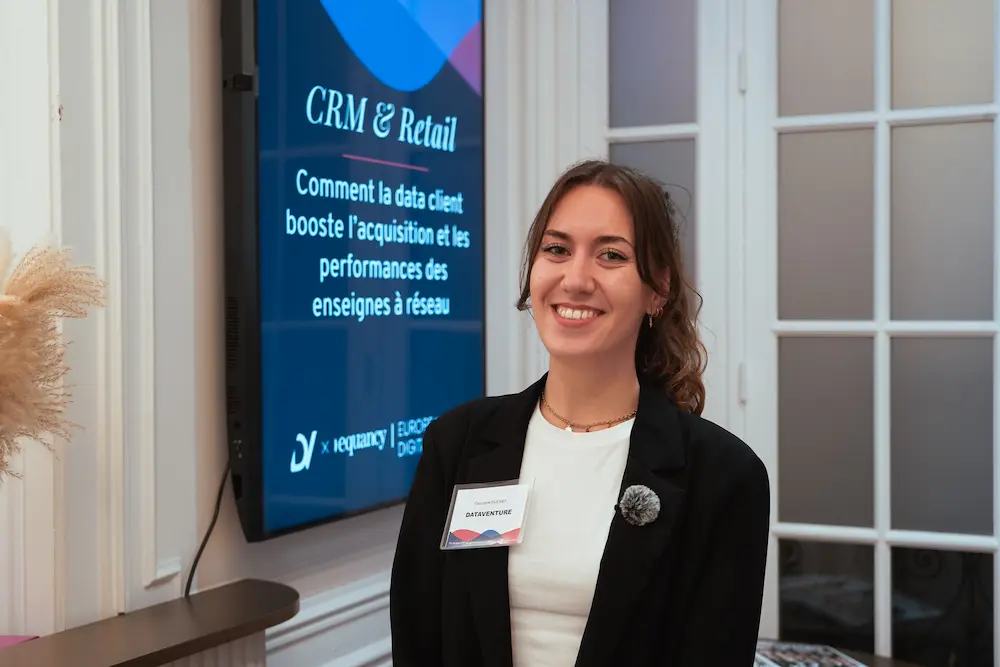 Smiling woman wearing a black blazer and white shirt standing indoors next to a screen displaying 'CRM & Retail' in French.