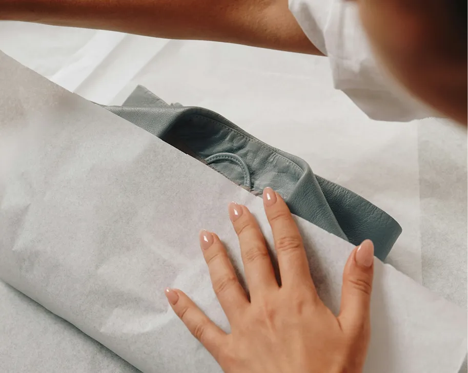 Person's hand with manicured nails wrapping a gray leather item in white tissue paper.