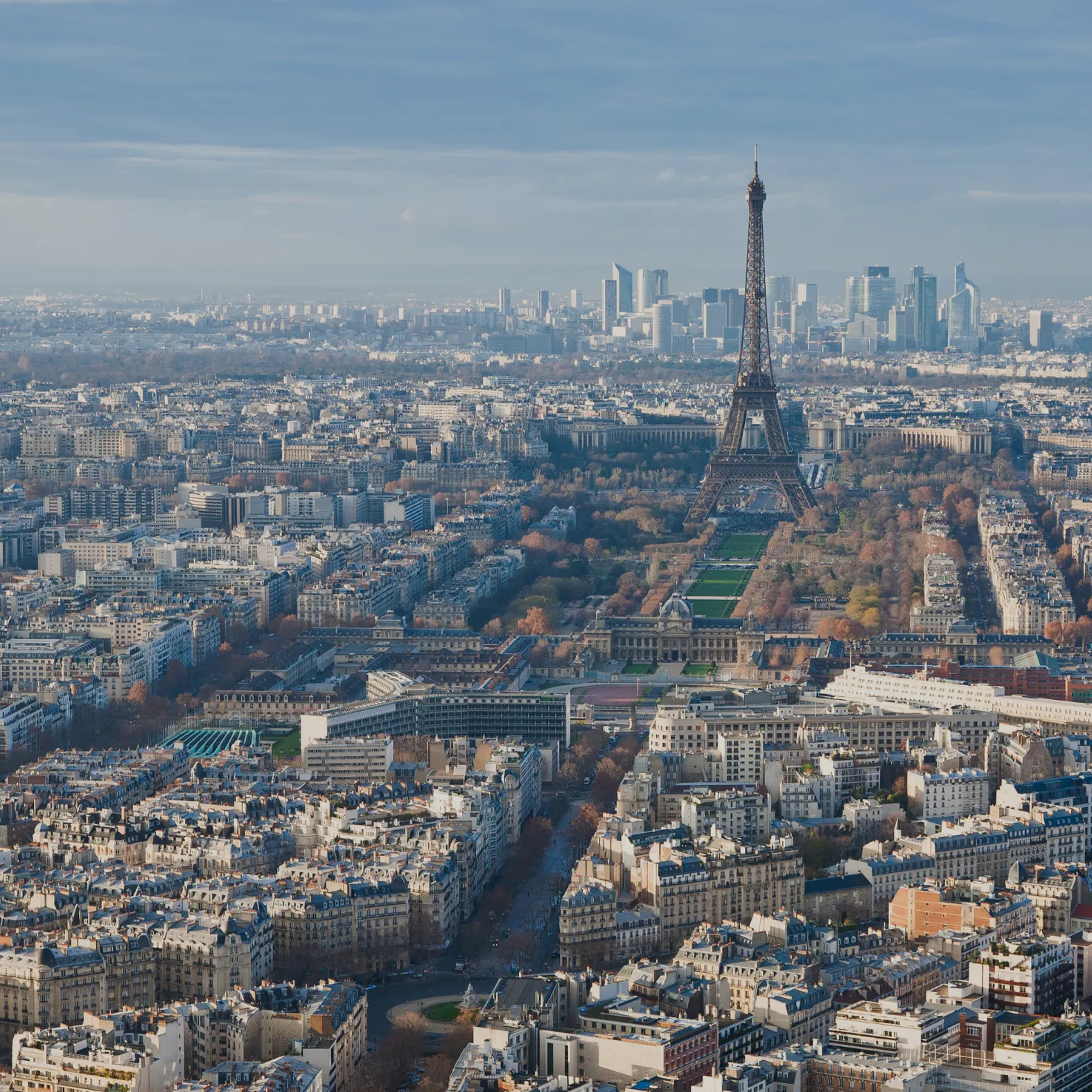 Aerial view of the Eiffel Tower surrounded by Paris buildings under a partly cloudy sky.