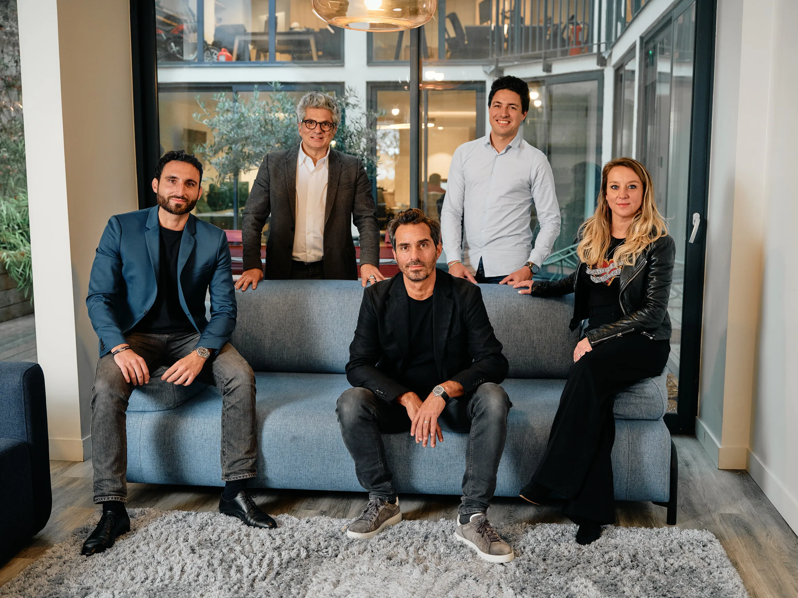 Five professionally dressed people posing in an office sitting and standing around a light blue couch.