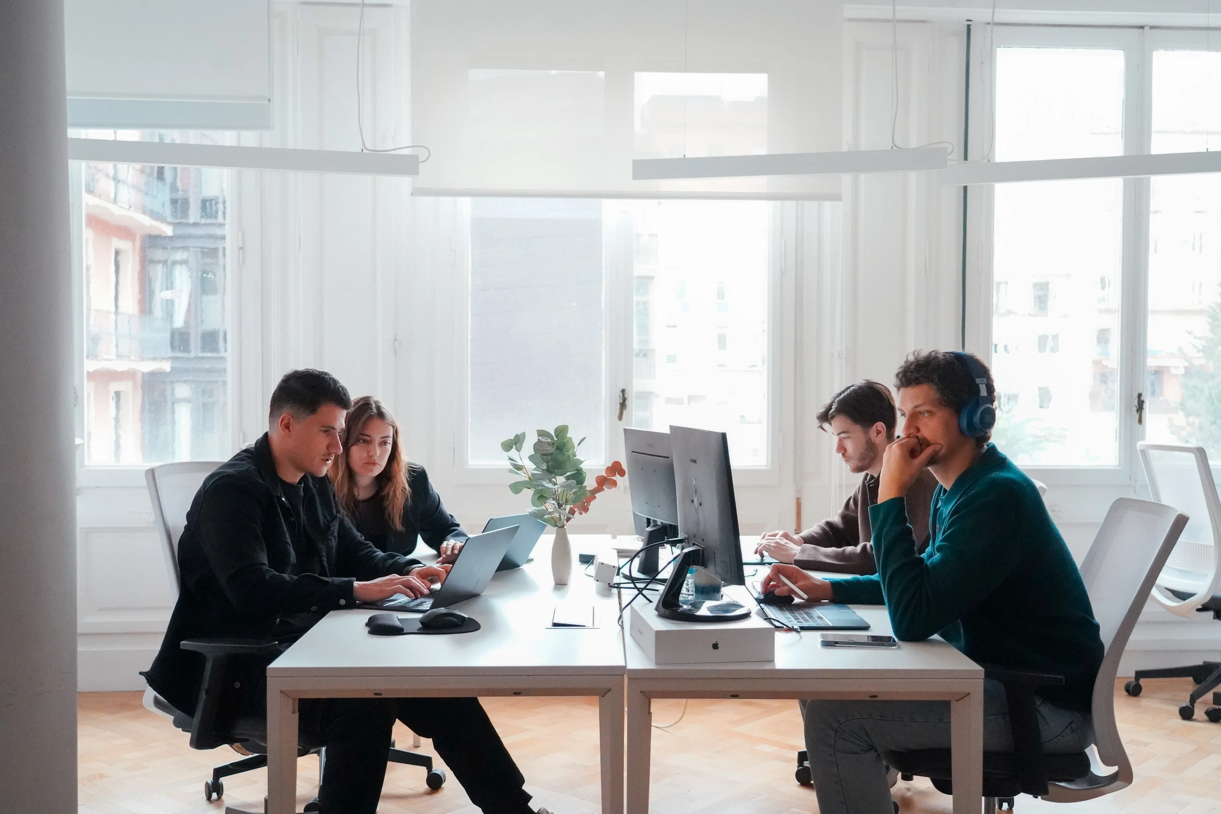 Four young professionals working at computers in a bright modern office with large windows.