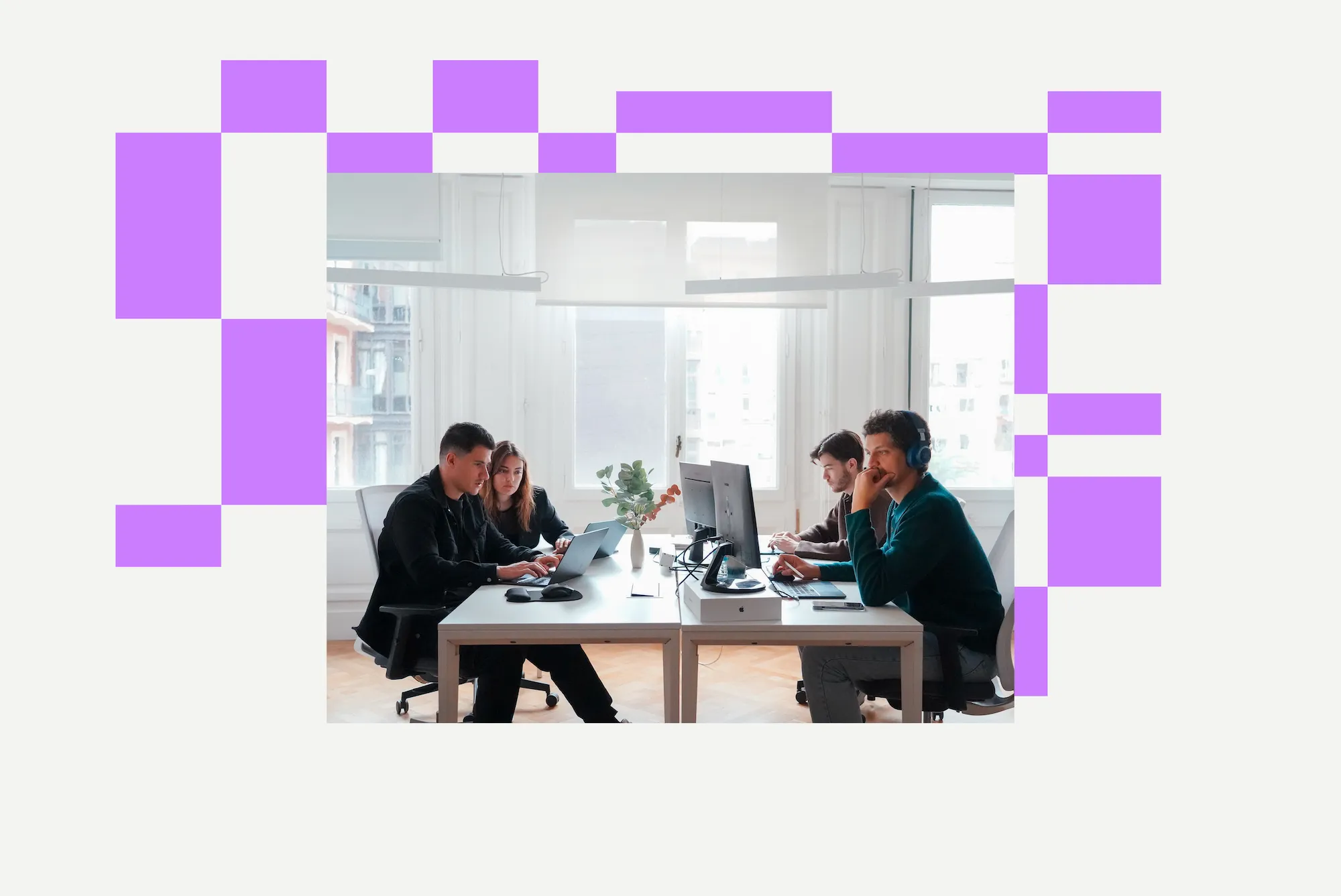 Four colleagues working in an office with laptops and desktop monitors at two desks facing each other by large windows.