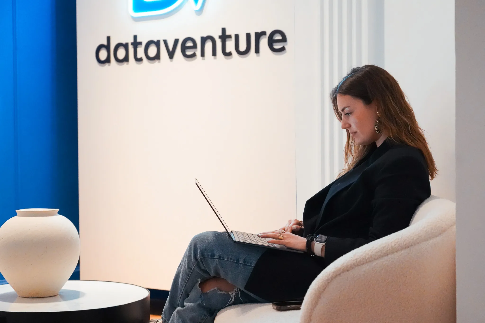 Woman sitting on a white chair using a laptop with a 'dataventure' sign in the background.