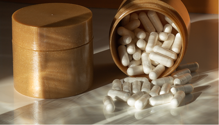 Biodegradable golden jar with white supplement capsules spilled on a reflective surface, showcasing sustainable packaging design in natural light.