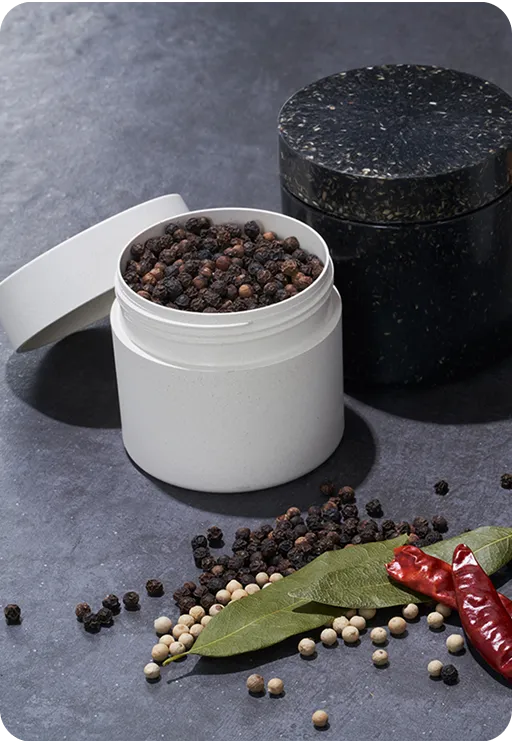 White spice jar filled with black peppercorns beside a black eco-friendly container, surrounded by dried chili peppers, bay leaves, and white peppercorns on a dark surface.