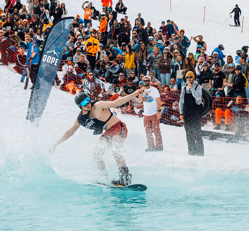 A crowd gathers to watch a man on a snowboard compete on a skim pool as he glides across the water