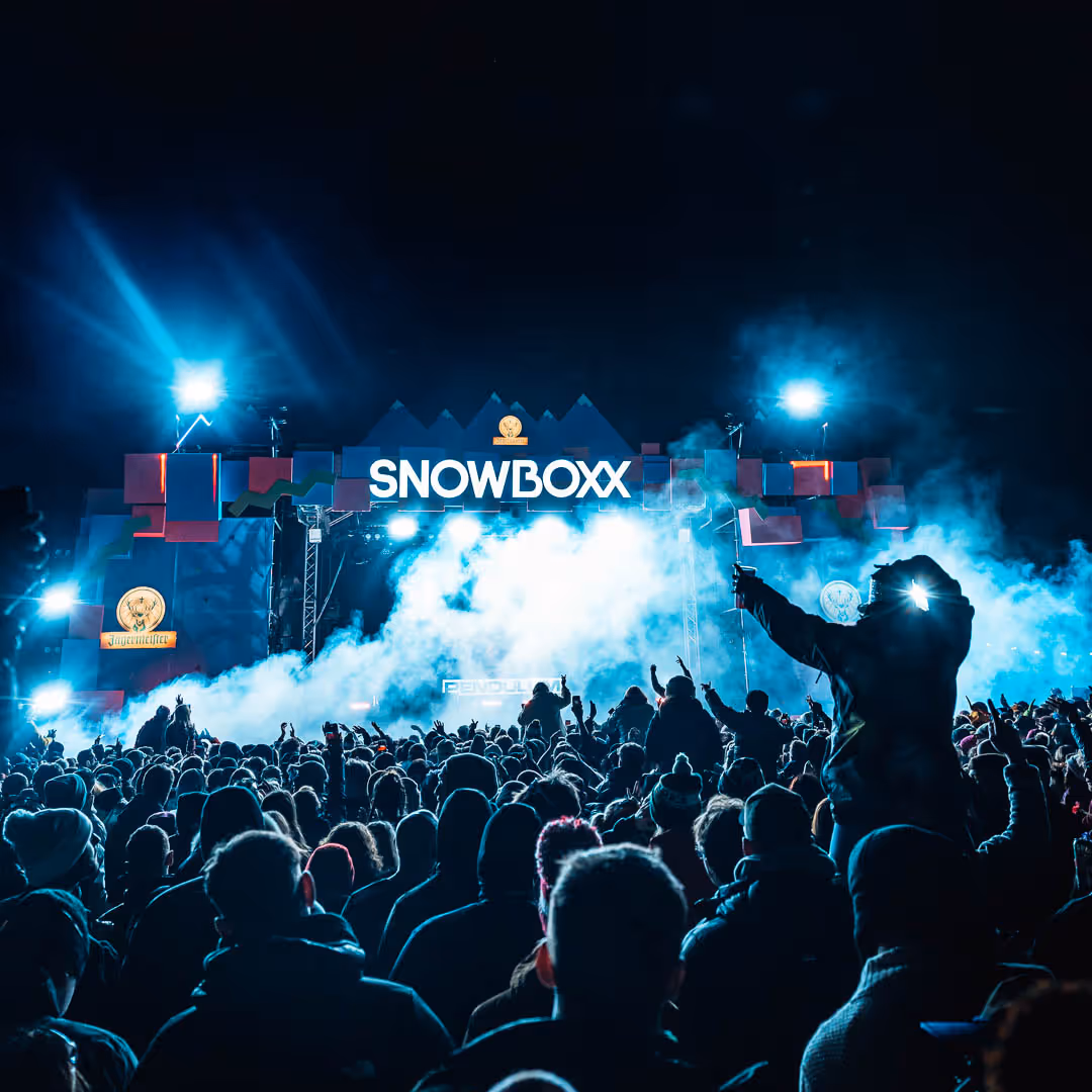 Large crowd shot overlooking the Snowboxx mainstage at night, silhouette of a girl on someone's shoulders, atmospheric smoke in front of the stage 