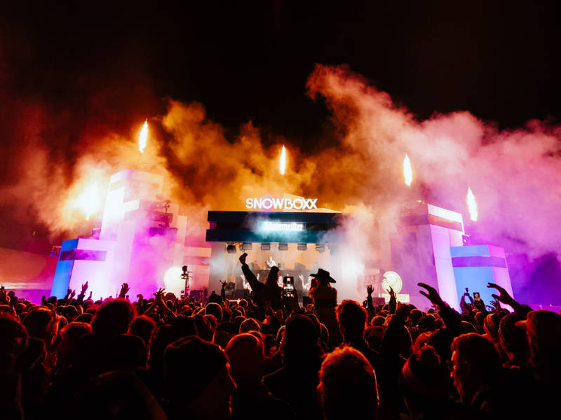 Large crowd shot overlooking the Snowboxx mainstage at night, silhouette of a girl on someone's shoulders, atmospheric smoke in front of the stage 